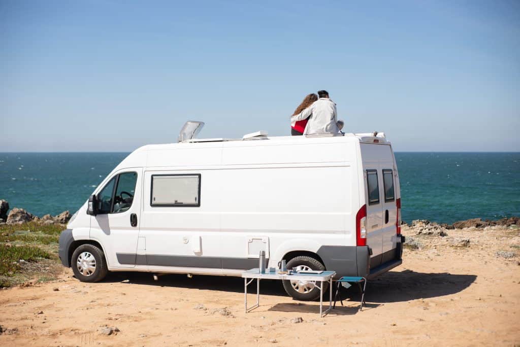 Back view of a couple sitting on top of a campervan on a beach
