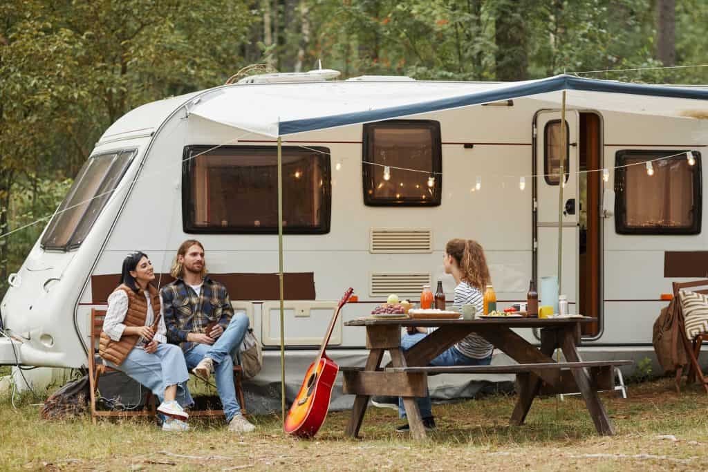 A group of adult friends sitting outside of a camper trailer with an awning at a picnic table