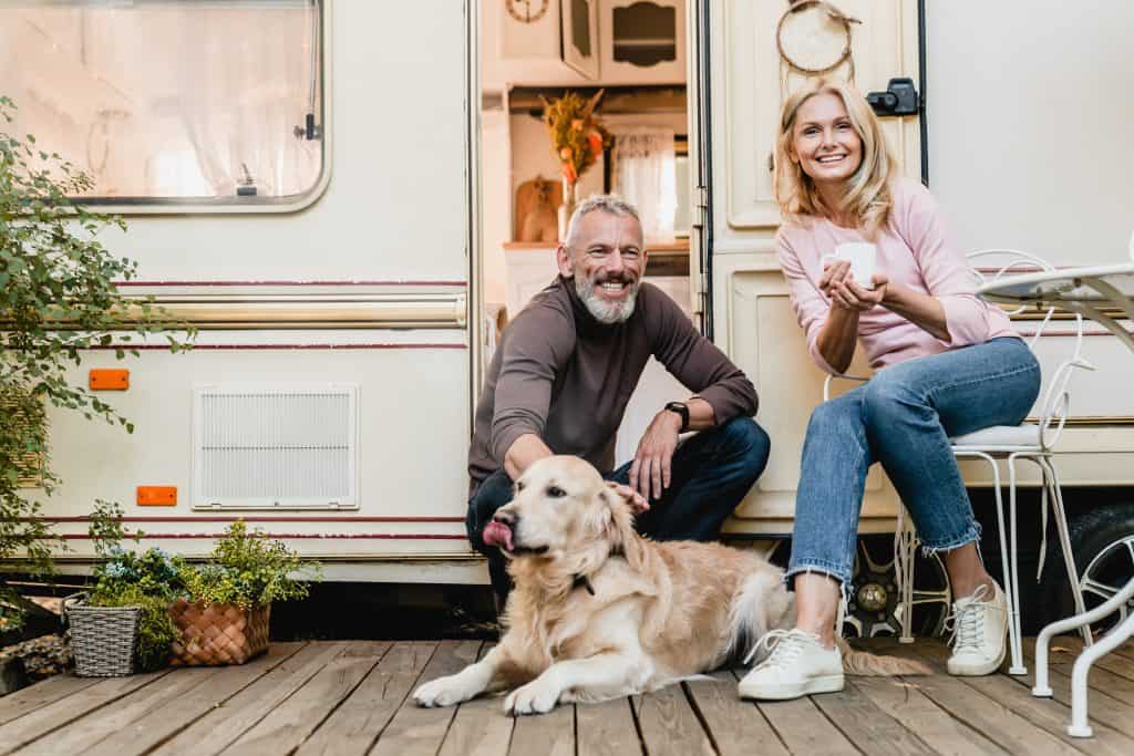 An elderly couple sitting outside of their camper with the door open and a dog lying on a deck